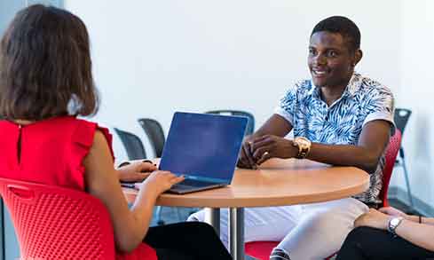 Two students work at a small table, one with a laptop open and typing.