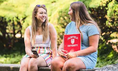 Student sitting on a stone wall smiling at each other on campus with a green backdrop.