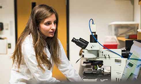 Student wearing a lab coat sitting in front of a microscope about to examine something.