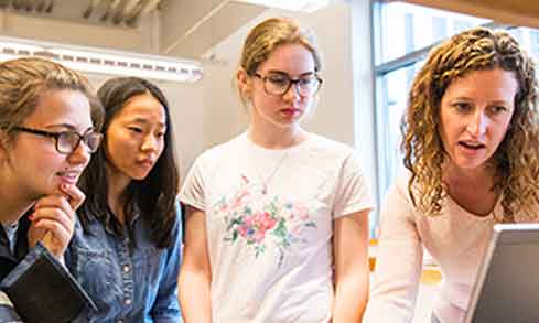 Three students and a professor look at an open laptop.