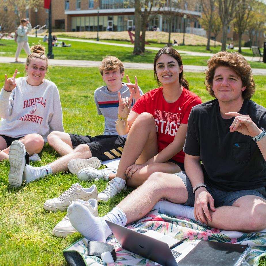 Four students sit outdoors in the grass on blankets holding up their hands to make a stag.