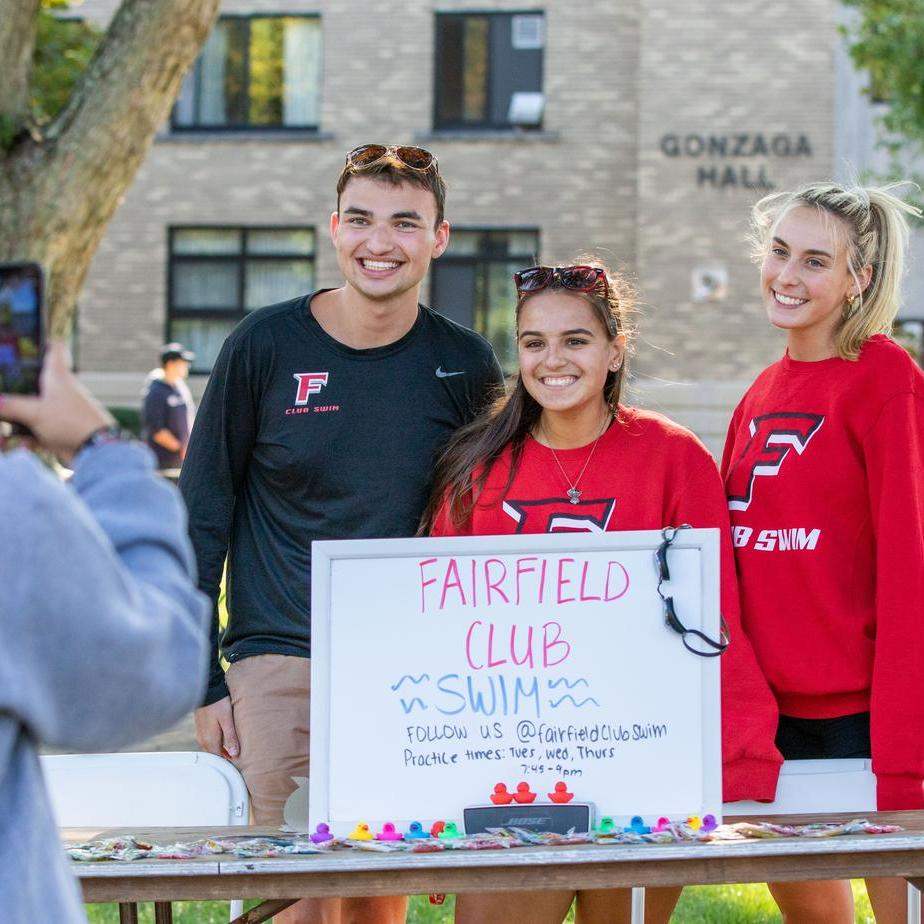 Three students pose for a photo behind a whiteboard that reads “Fairfield Club Swim, Follow Us @fairfieldclubswim”.