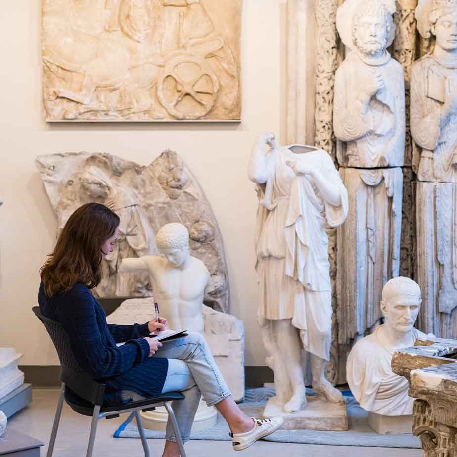 A woman seated in a chair, surrounded by an extensive array of statues in a well-lit space.