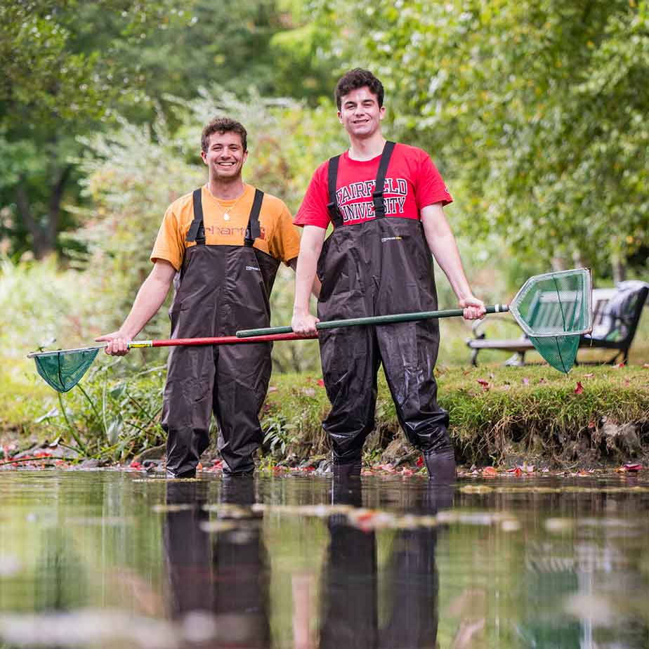 Two men in overalls stand in a pond, each holding a net, enjoying a day of fish lifecycle studies in Bellarmine Pond.