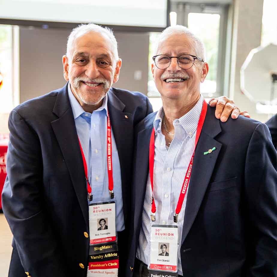 Two older friends in suits smile and shake hands at a reunion event.