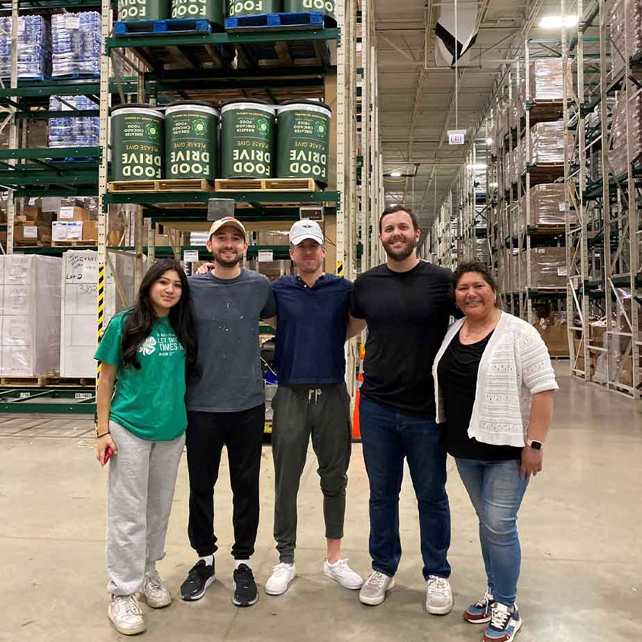 Four individuals pose in front of a warehouse stocked with numerous cans, showcasing a vibrant industrial setting.