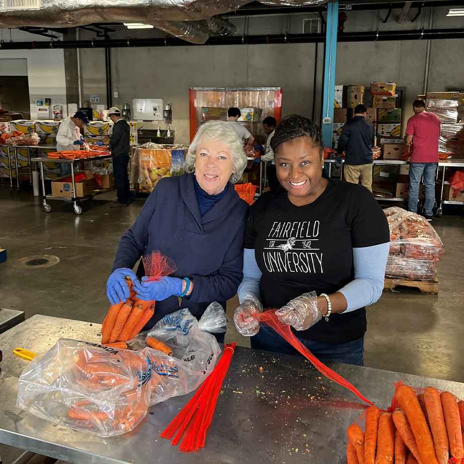 two women cut carrots and smile at the camera. one is wearing a Fairfield University shirt.