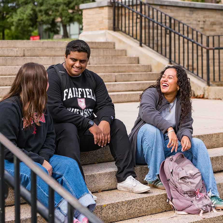 Three students seated on the steps of a building, engaged in conversation and enjoying their time together.