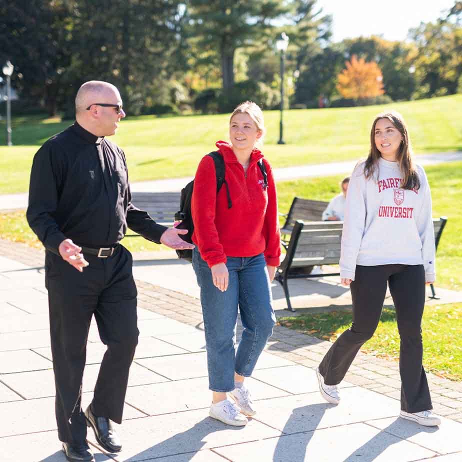 Three individuals stroll along a sidewalk, featuring a jesuit priest and two women.