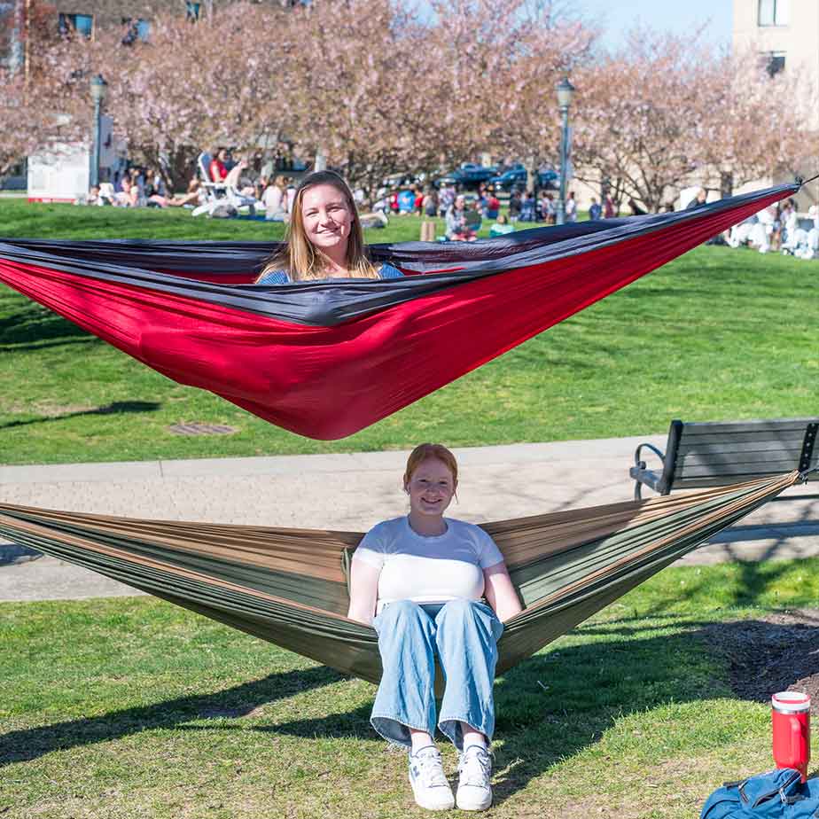 Two women relax in a hammock, surrounded by a lush grassy field under a clear blue sky.