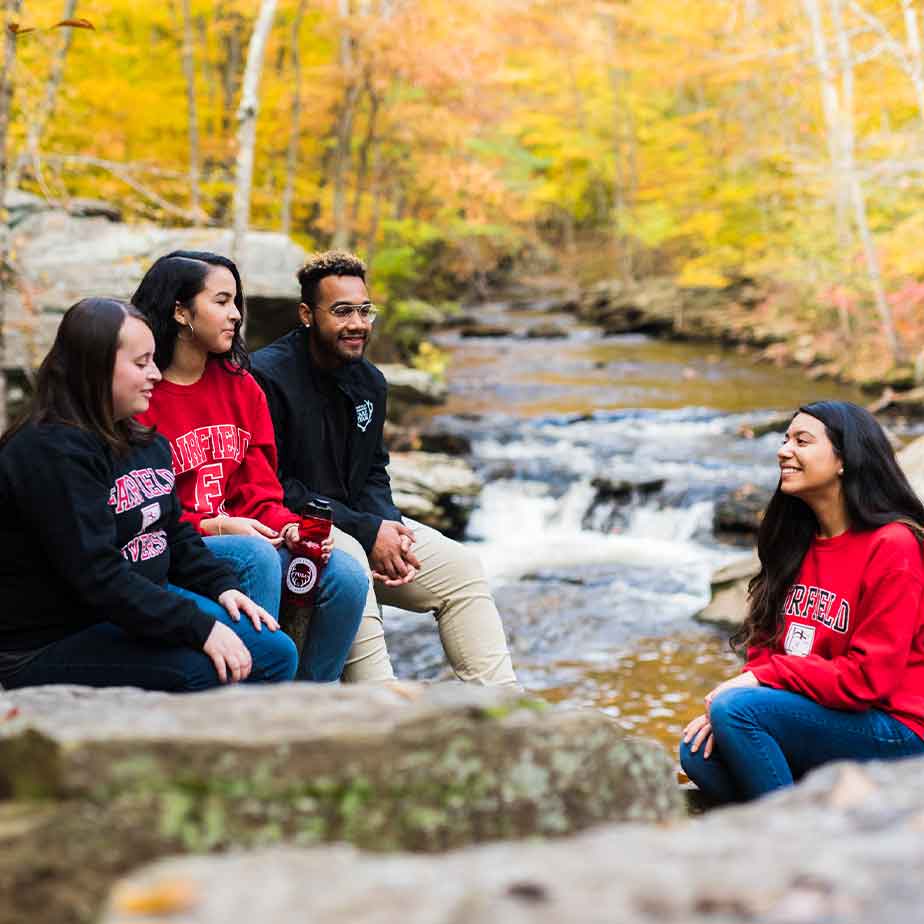 Four students seated on rocks beside a flowing stream, engaged in conversation and enjoying the natural surroundings.