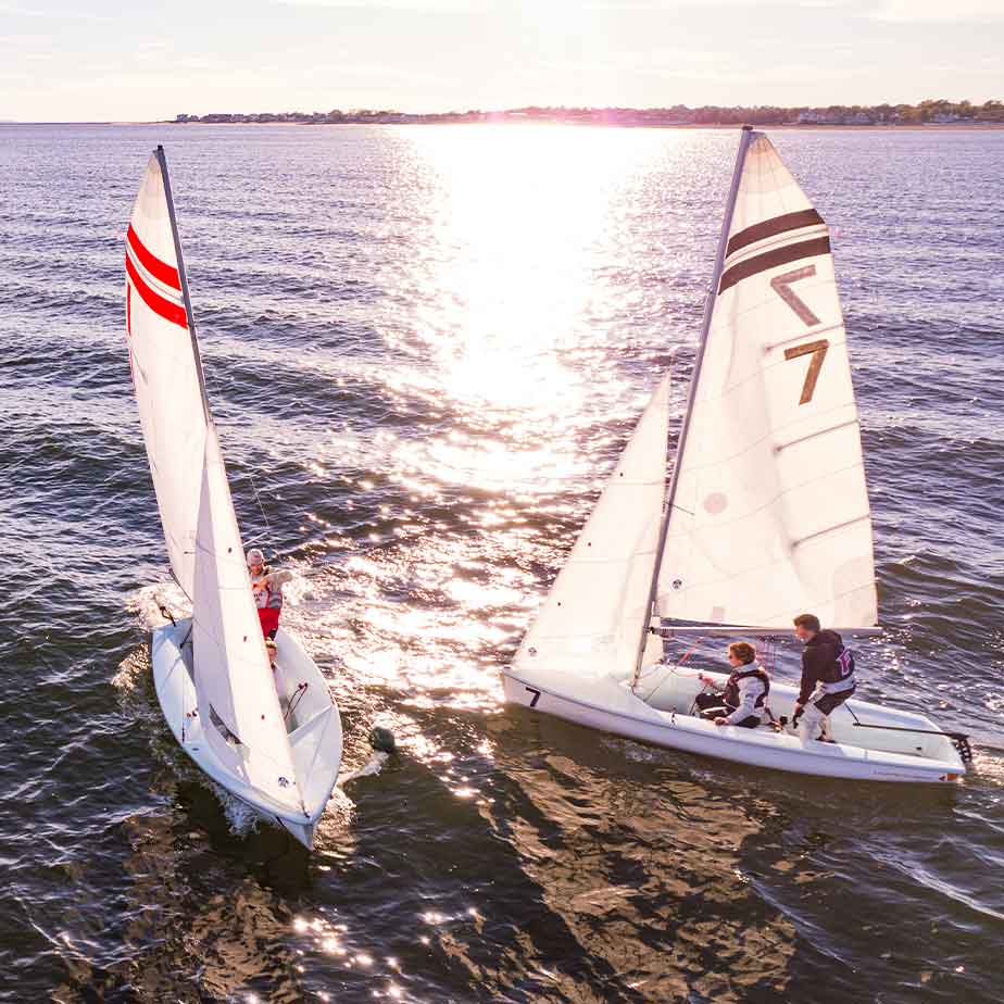 Two sailors maneuver a sailboat on the open ocean, surrounded by water and a bright sky.