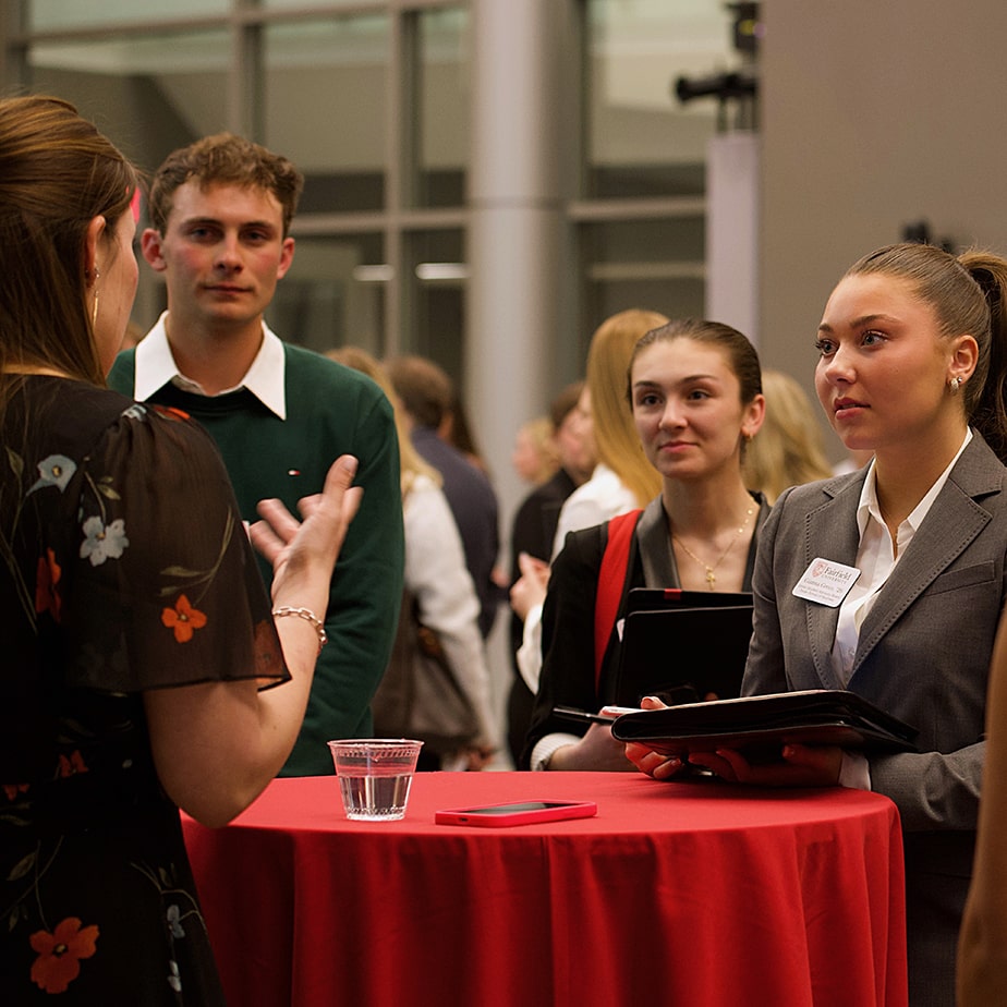 Students wearing formal attire standing around a table at an event listening to a speaker.