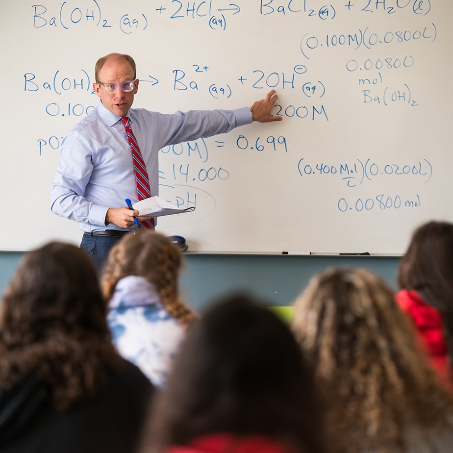 Professor stands at the front of a classroom and is explaining written equations on a whiteboard.