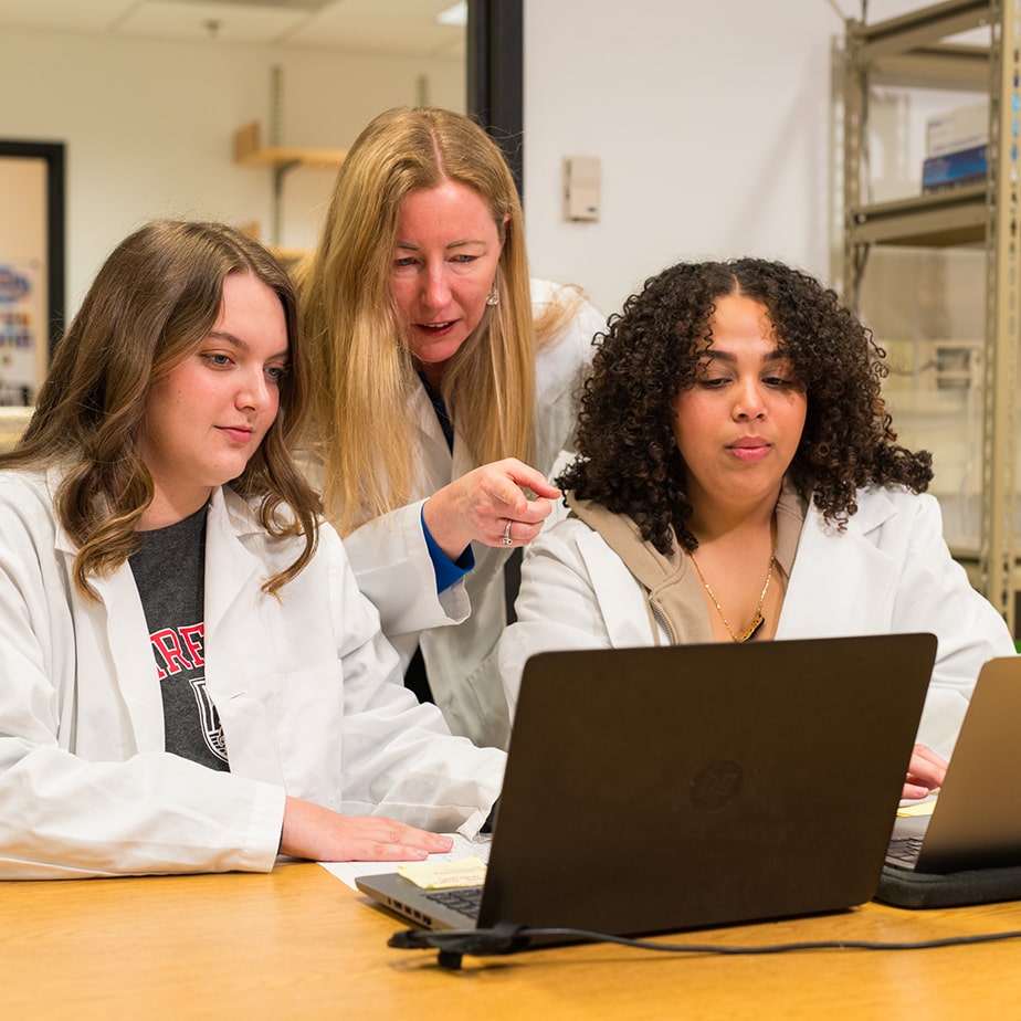 Students wearing lab coats have their teacher explain something to them as she points at their computers.