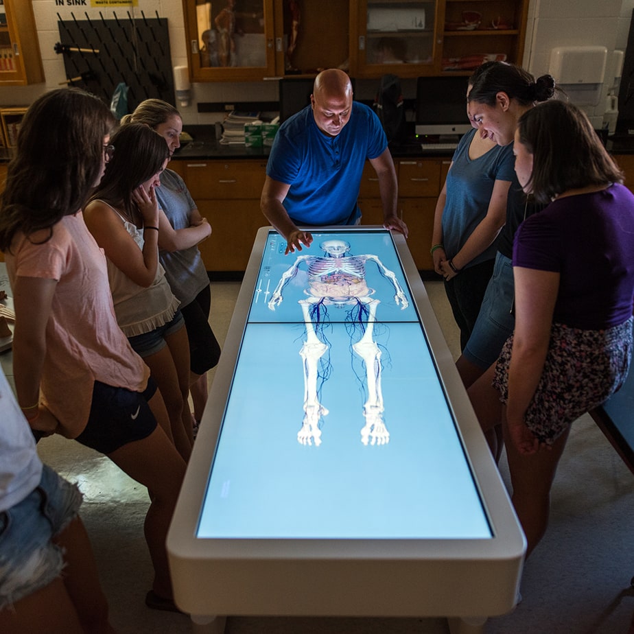 Students stand around an illuminated screen the size of a coffin and listen to their professor explain something while pointing to a virtual skelton on the screen.