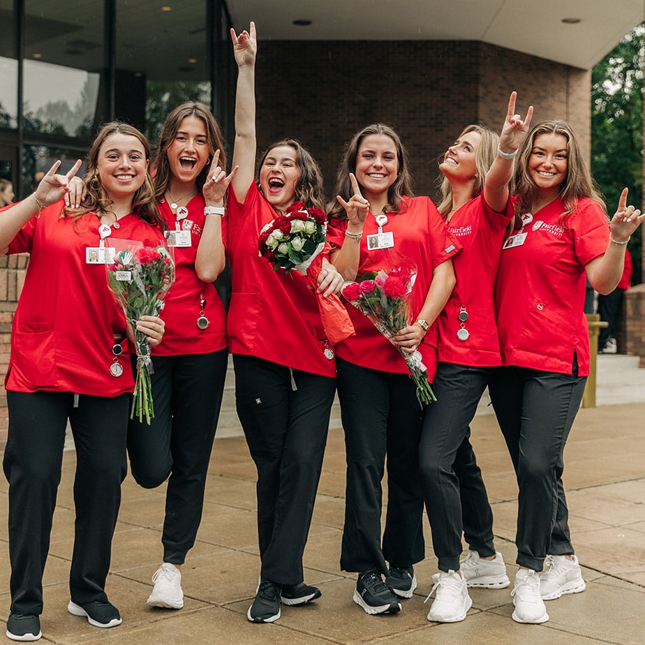 Egan nursing students posing for a photo wearing their red Fairfield scrubs and holding flowers while doing stag hand signs.