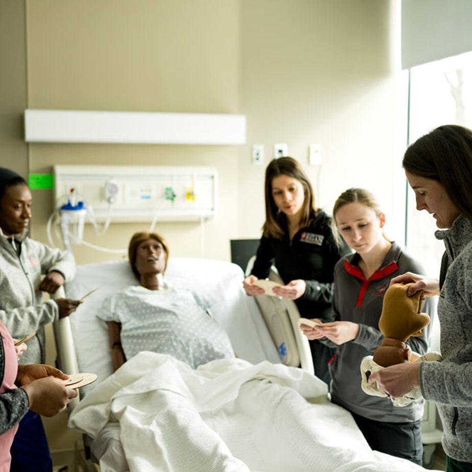 Egan students watching a demonstration done by their professor in a room with a simulation doll in a hospital bed.