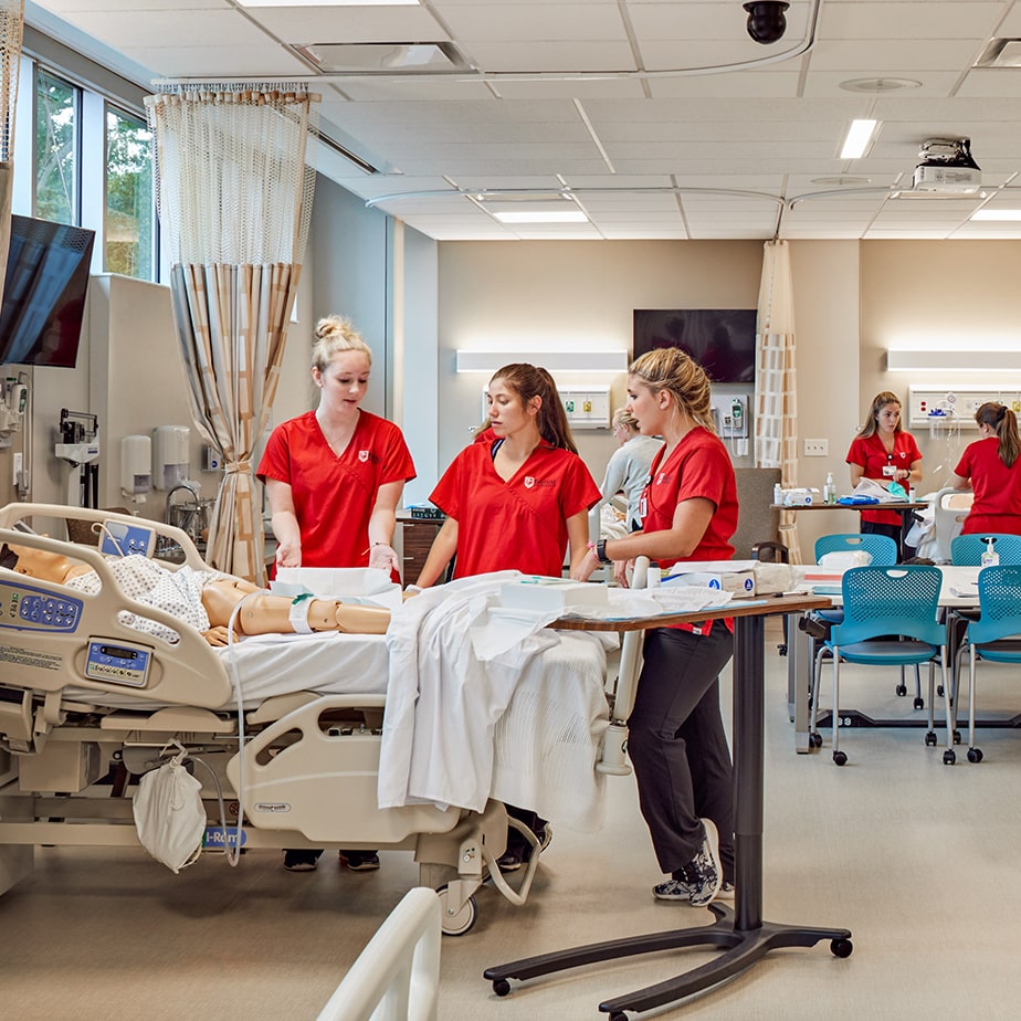 A mock hospital room full of Egan nursing students wearing red Fairfield scrubs and tending to dummies.