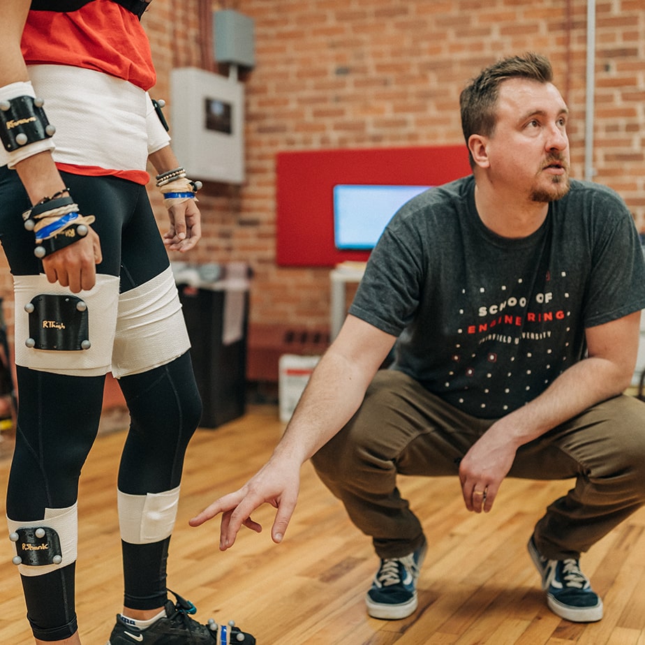 Professor pointing to the leg of a student wearing an engineering suit. 