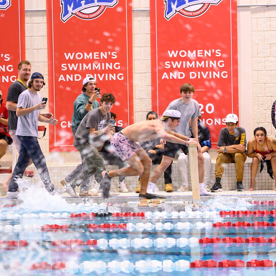 Student speeds across the university pool on a man made flotation device while his peers watch and cheer for him outside of the pool. Banners in the background say “Women’s swimming and diving”. 