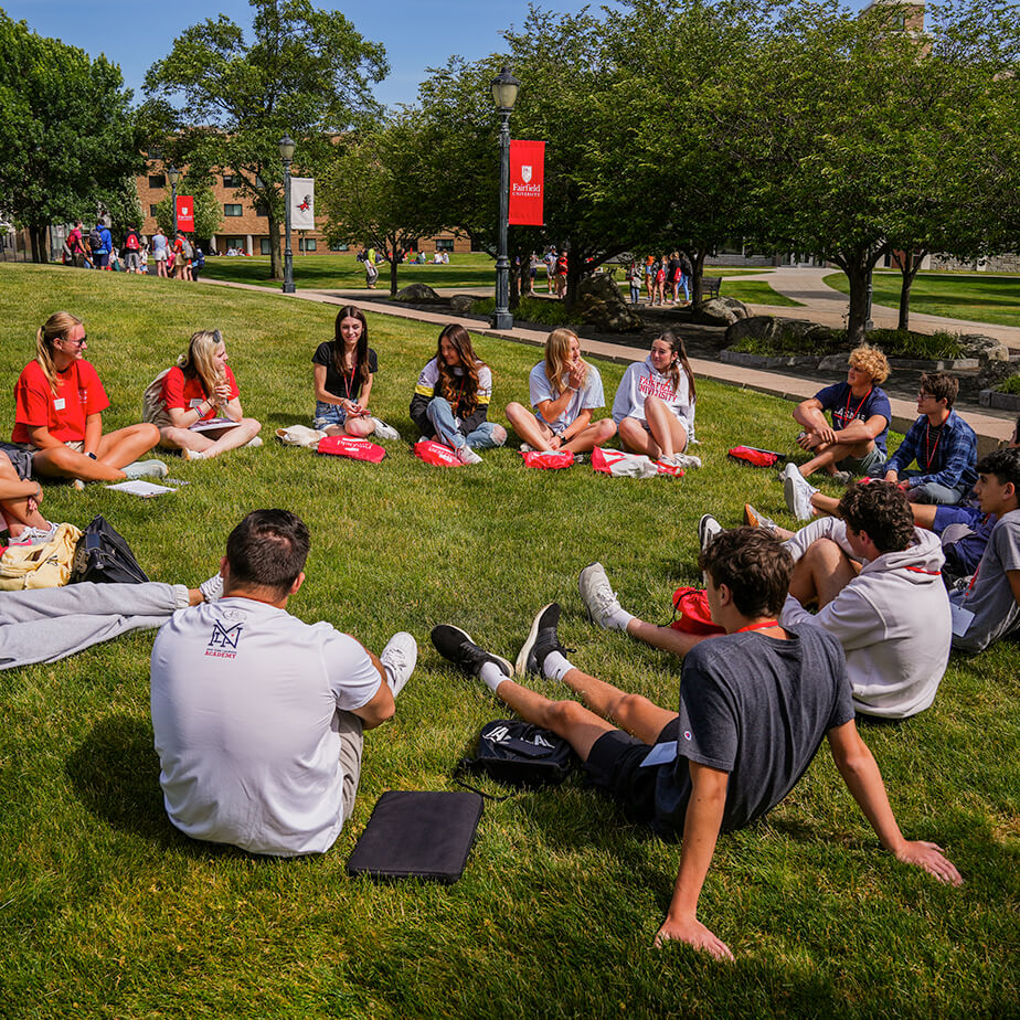 A gathering of new students sitting on the grass, engaged in conversation and enjoying the pleasant outdoor environment.