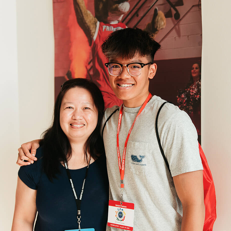 A young man and woman stand side by side, smiling, in a casual indoor setting. 