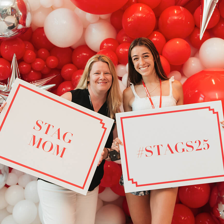 Two women proudly display signs reading "Stag Mom" and "#Stags25" showcasing their support and celebration of joining the newsUniversity community.