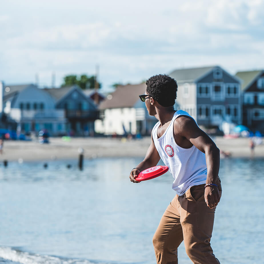 A student joyfully throws a frisbee on a sunny beach, with waves gently lapping at the shore in the background.