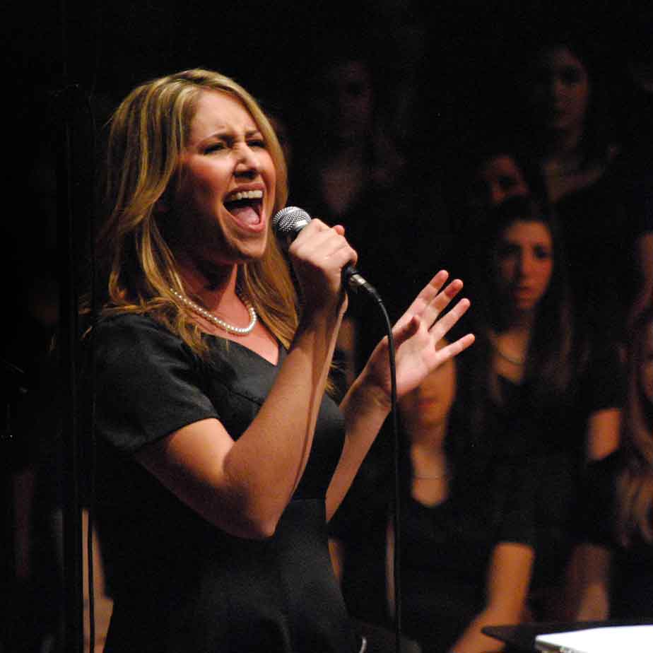 A woman passionately sings into a microphone, surrounded by a choir harmonizing behind her.