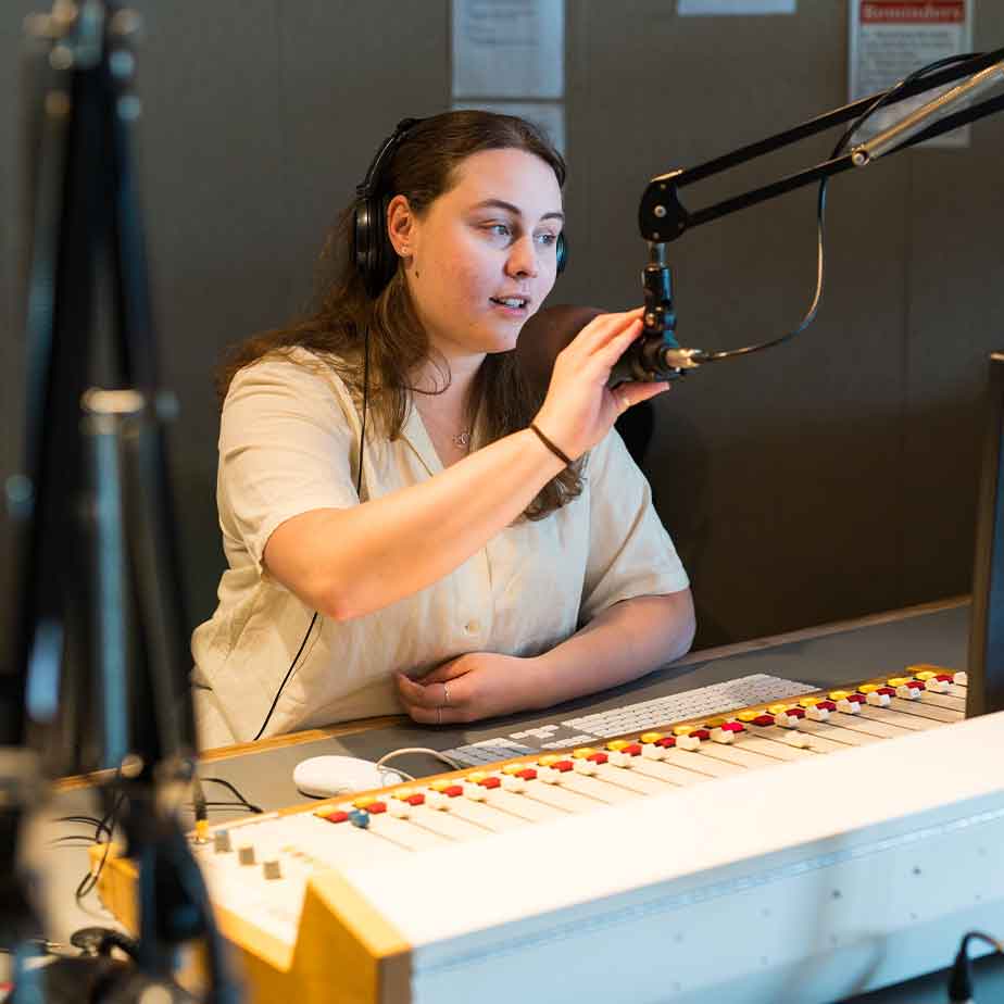 A woman speaking into a microphone in a radio studio, surrounded by sound equipment and acoustic panels.