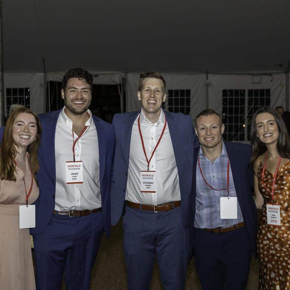 Five men and two women pose standing together at an evening alumni event in an outdoor tent.