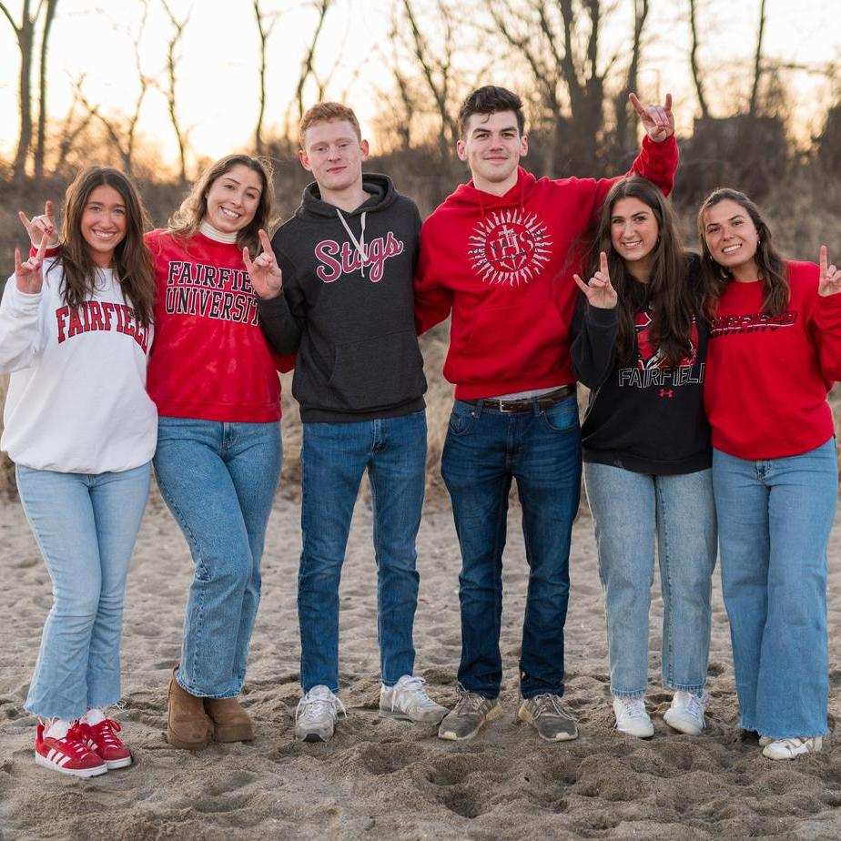 Six Fairfield students pose on the beach with trees behind them, giving the "stag" sign with their hands.