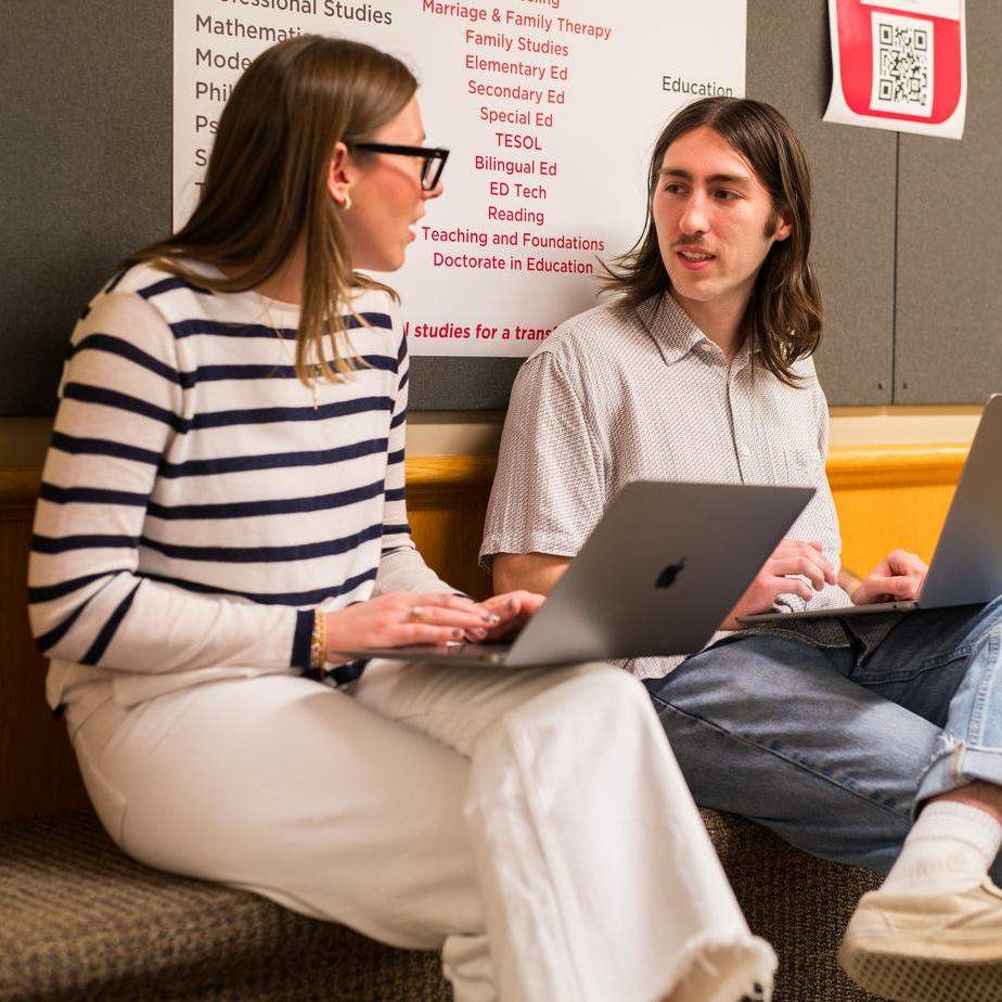 A student and professor sit with laptops and chat in an academic building. 