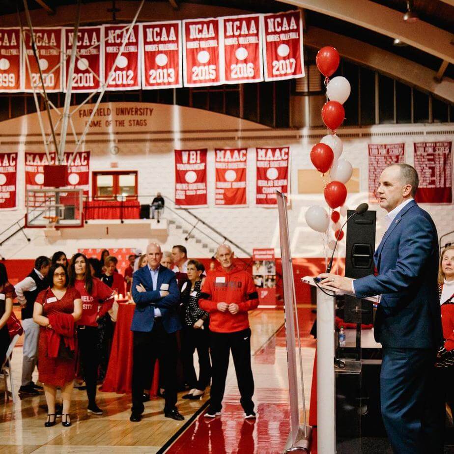 A man in a suit speaks into a microphone at a podium in front of red and white balloons to a crowd of people.