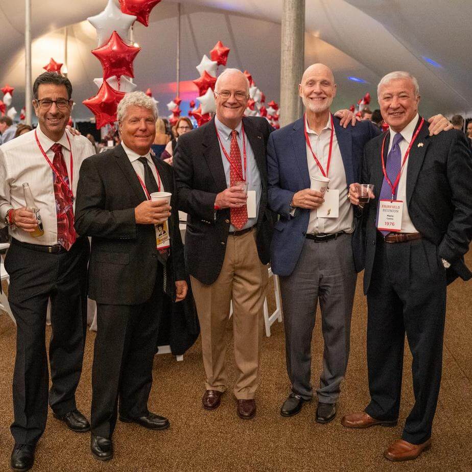 A group of alumni in suits pose in front of red and white star-shaped balloons.