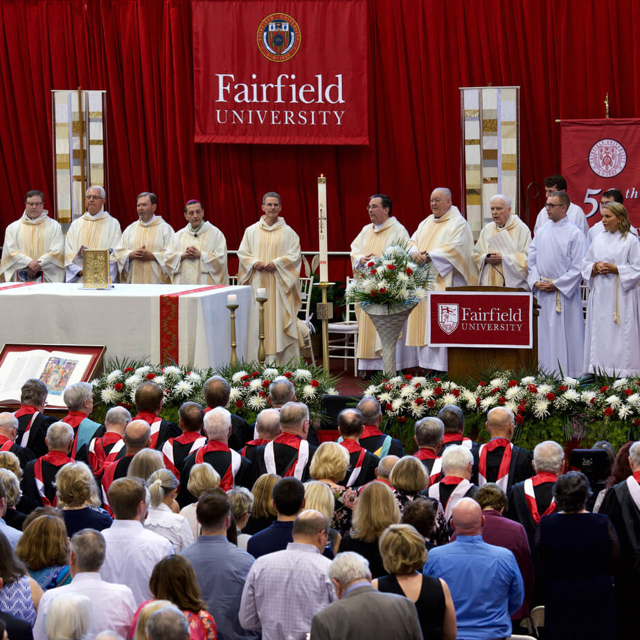 A crowd of alumni stand in front of a stage full of priests with signs that read “Fairfield University”.