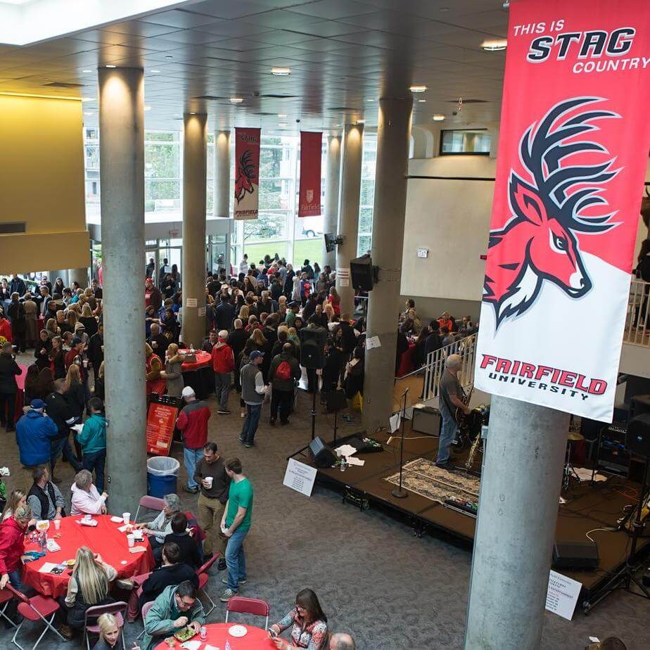 A crowd of people fill a large interior below a sign that reads “This is Stag Country; Fairfield University”.