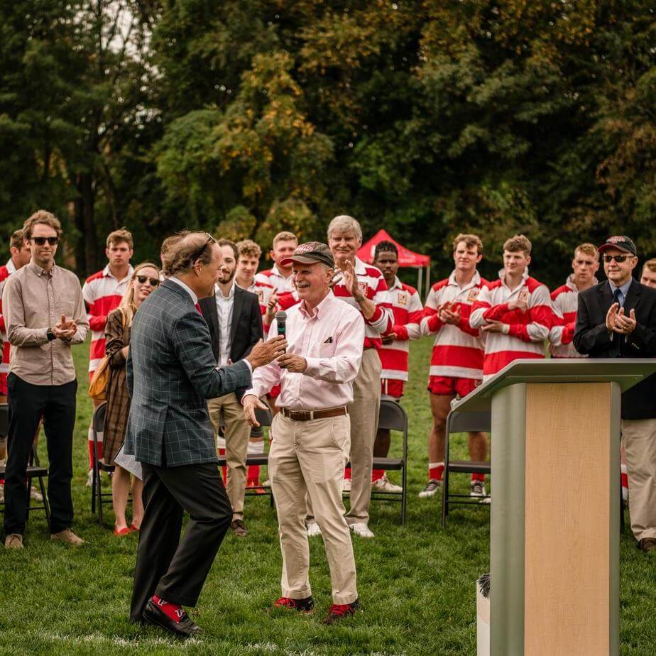 Two people shake hands in front of a large crowd of onlookers.