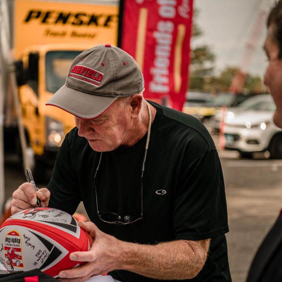 A man in a gray Fairfield hat signs a rugby ball with a black sharpie.