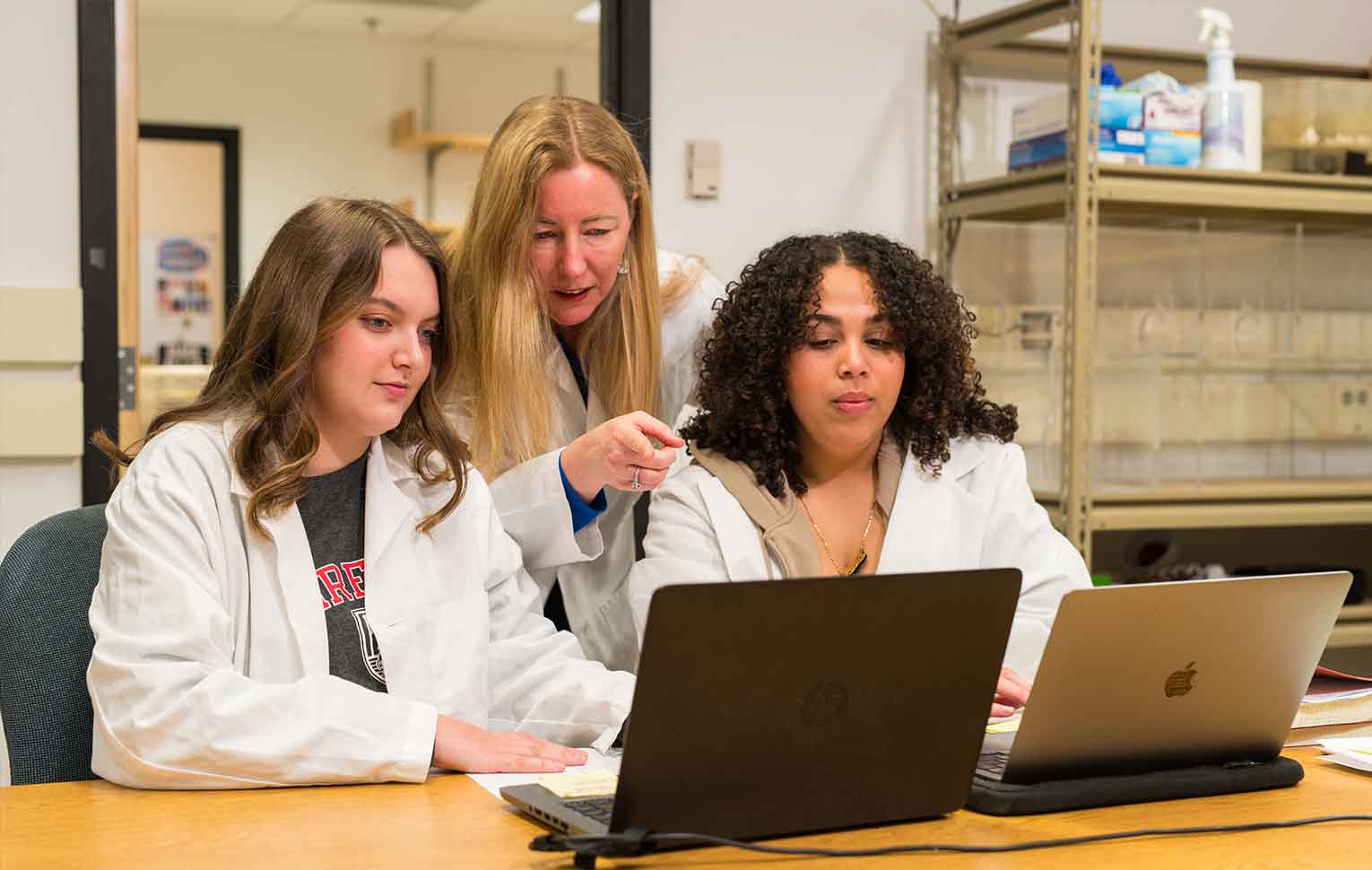 two students in white lab coats work on laptops with a professor