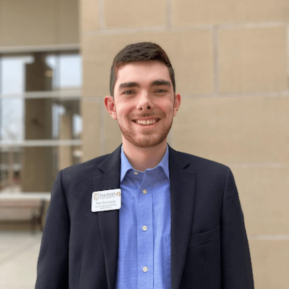 Max Berryman smiles in a blue button-up shirt in front of an academic building.