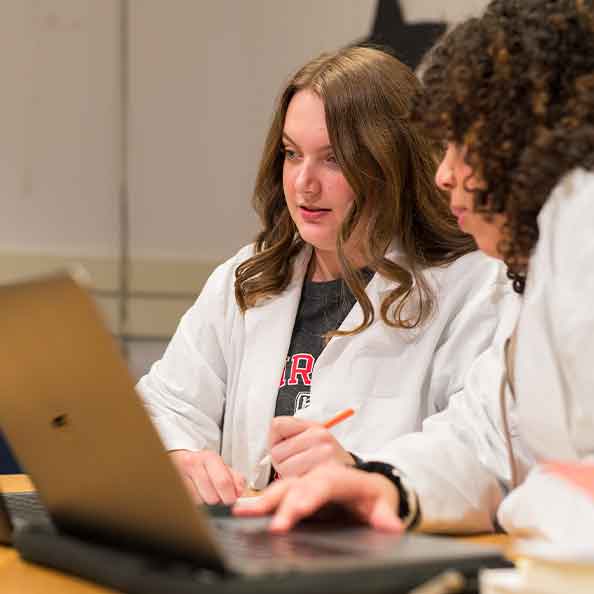 Two female scientists in lab coats engaged in a discussion while working on a laptop in a lab environment.