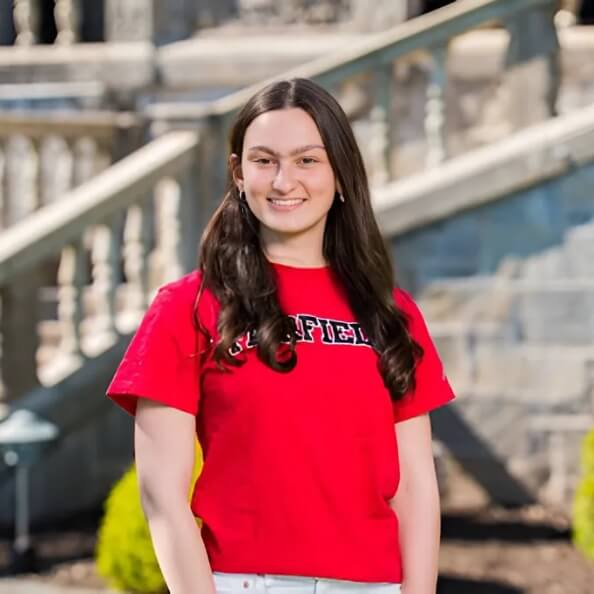 Student smiling and looking at the camera head on in front of an older stone building on campus. She is wearing a red Fairfield shirt. 