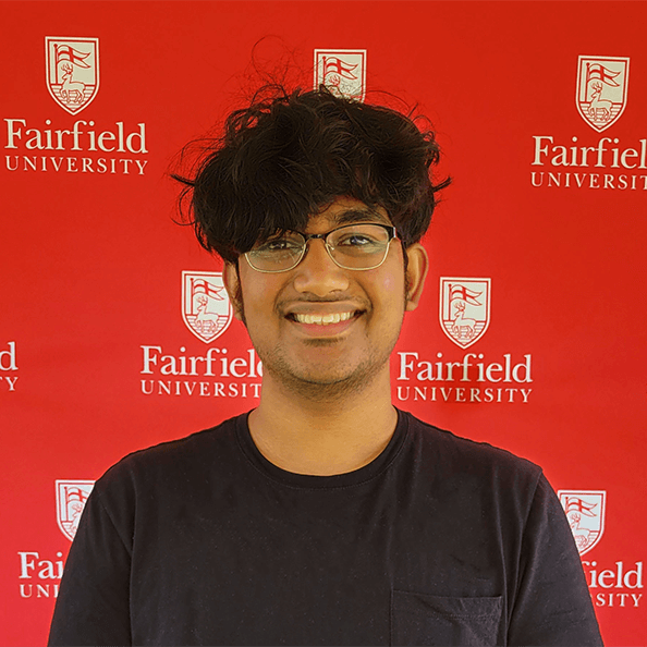 Nahush smiles in front of a red Ƶ University background.