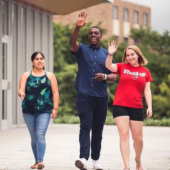 three students walking across campus and waving at friends.