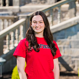 A student poses in front of an older stone building on campus. She is wearing a red Fairfield shirt.