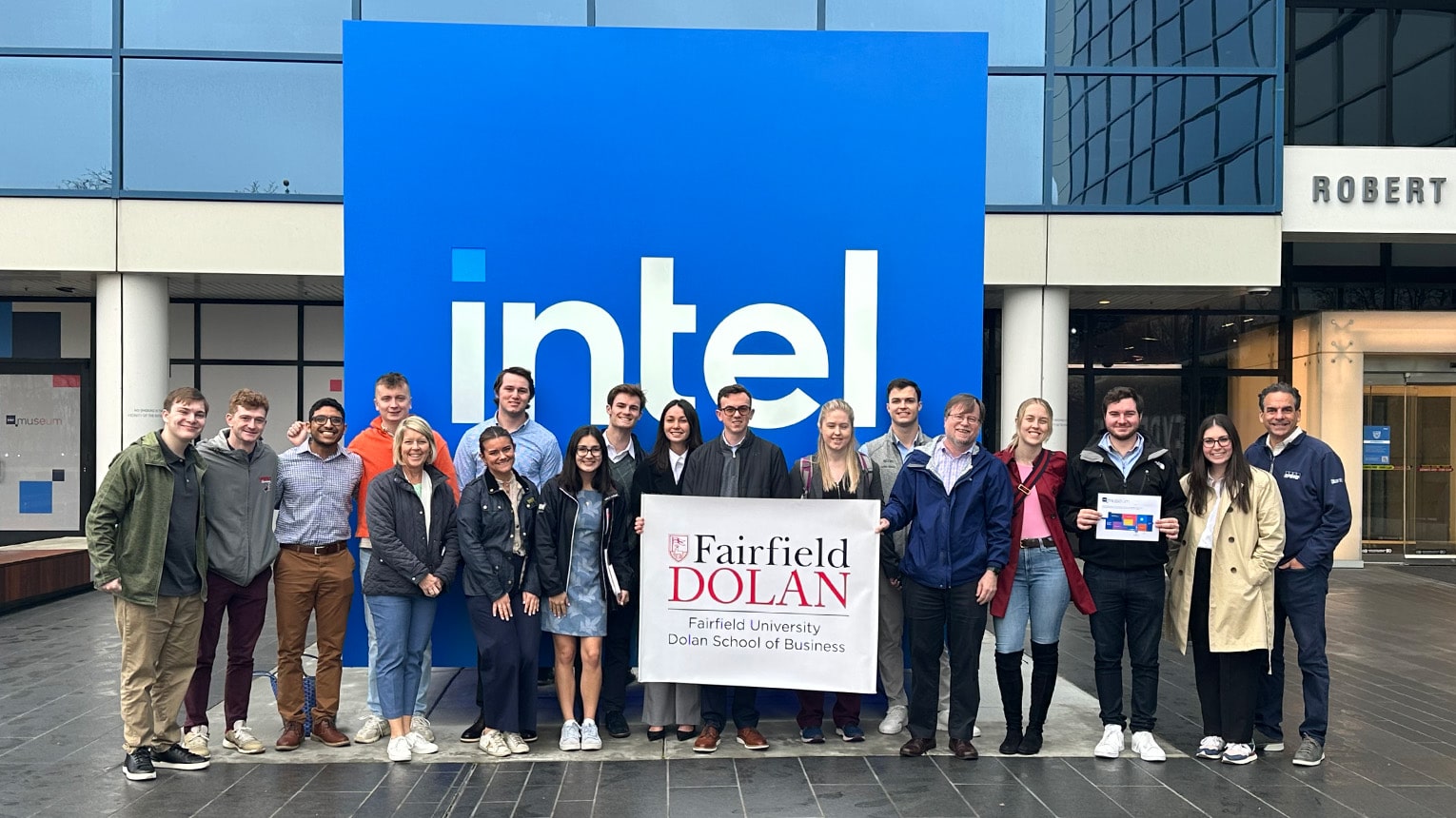 Students holding a Fairfield Dolan sign while grouped and lined up in front of the exterior, blue Intel building marker.