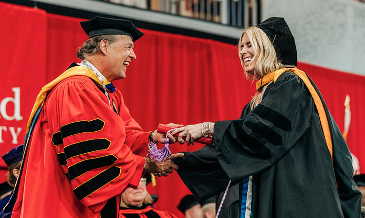 Mark Nemec in red commencement attire shaking a students hand at Commencement.