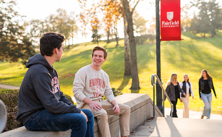 Two students sitting together on a bench, with a Fairfield University banner prominently displayed overhead.